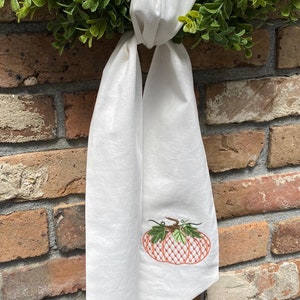 May include: A white fabric towel featuring an embroidered orange pumpkin with a cross-hatched pattern and green leaves. The towel is tied in a knot at the top and hangs vertically, displaying the detailed stitching of the pumpkin design.