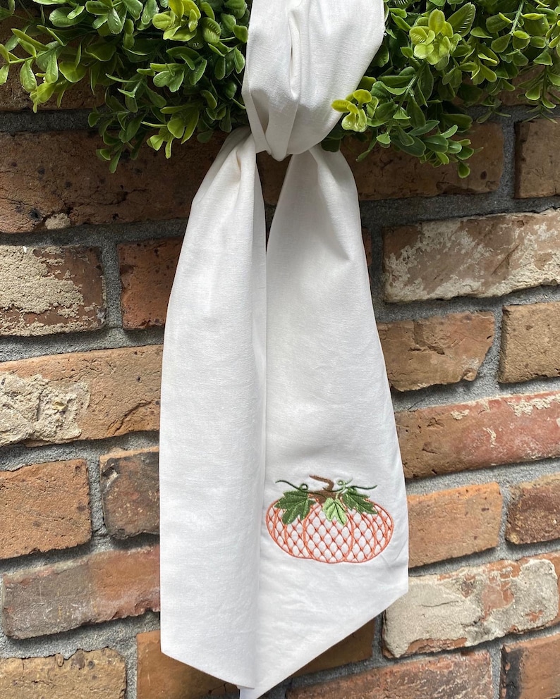 May include: A white fabric towel featuring an embroidered orange pumpkin with a cross-hatched pattern and green leaves. The towel is tied in a knot at the top and hangs vertically, displaying the detailed stitching of the pumpkin design.