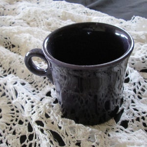 May include: A purple ceramic coffee mug with a rounded handle sits on a white crocheted doily.
