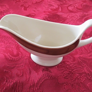 以下が含まれることがあります： 茶色のストライプ模様が入った白いセラミック製のグレイビーボート。グレイビーボートには、注ぎやすいようにハンドルと注ぎ口が付いています。赤い模様のテーブルクロスの上に置かれています。
