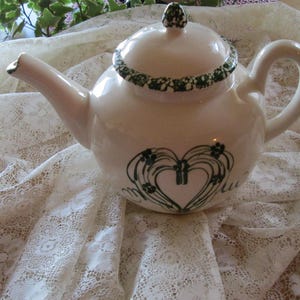 Könnte beinhalten: Cremefarbene Keramik-Teekanne mit Deckel und Henkel. Die Teekanne hat ein grünes Blumendesign am Rand und ein herzförmiges Blumendesign auf der Vorderseite. Sie steht auf einer weißen Spitzentischdecke.