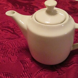 Könnte beinhalten: Eine weiße Teekanne aus Keramik mit Deckel und Henkel. Die Teekanne steht auf einer roten Tischdecke mit Blumenmuster.