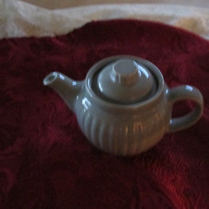 May include: A white ceramic teapot with a lid and a ribbed design. The teapot is sitting on a red fabric surface.