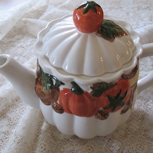 May include: White ceramic teapot with a lid decorated with a tomato, pumpkins, and leaves. The teapot has a handle and a spout.
