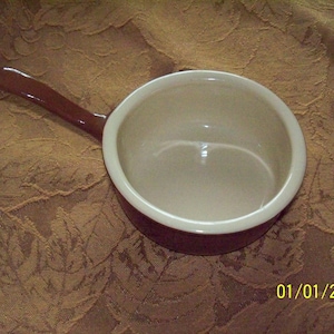 以下が含まれることがあります： クリーム色の内側と茶色の取っ手を持つ、小さな茶色のセラミック製のラメイクイン。ラメイクインは茶色の布の表面の上に置かれています