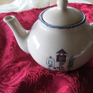 May include: A white ceramic teapot with a blue trim and a birdhouse design. The teapot is sitting on a red tablecloth.