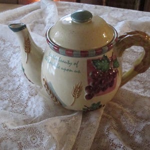 Puede incluir: Una tetera de cerámica con un diseño marrón claro, crema y verde. La tetera tiene una tapa con la parte superior azul y un asa con un diseño marrón y crema. La tetera tiene una cita en el lateral que dice "La pequeña belleza de la vida está sobre nosotros."