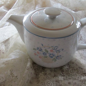 Peut inclure: Théière en céramique blanche avec un motif floral et un couvercle. La théière a une bordure brune et un motif floral bleu et blanc avec un panier de fleurs.