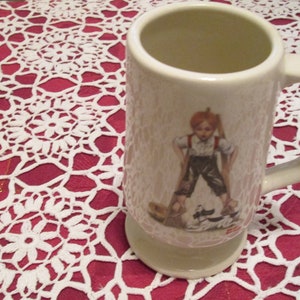 Puede incluir: Taza de cerámica color crema con asa, con una superficie texturizada y una ilustración impresa de un niño. El niño lleva tirantes y está de pie sobre un pequeño barco. La taza está sobre una superficie roja con un patrón de encaje blanco.