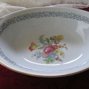 May include: Oval white serving bowl with a floral design in the center. The bowl has a blue floral border and gold trim. The handles are also trimmed in gold. The floral arrangement includes pink, yellow, orange, and blue flowers.