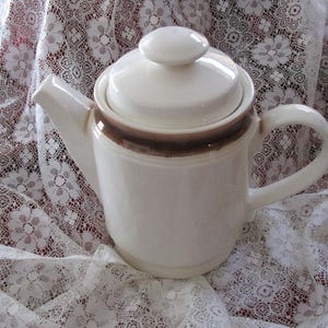 Puede incluir: Una tetera de cerámica blanca con una banda marrón alrededor del centro. La tetera tiene una tapa y un asa. Está sentada sobre un mantel de encaje blanco.