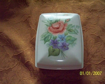 May include: A white ceramic trinket box with a floral design. The box features a pink rose with green leaves and purple flowers.