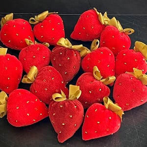 May include: A collection of 14 red velvet strawberry pincushions with gold trim and sequins. The pincushions are arranged in a heart shape.