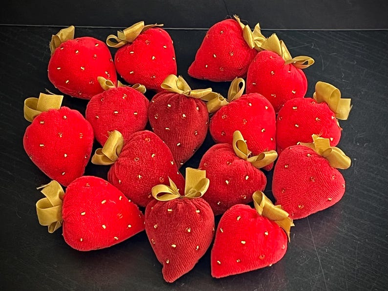 May include: A collection of 14 red velvet strawberry pincushions with gold trim and sequins. The pincushions are arranged in a heart shape.