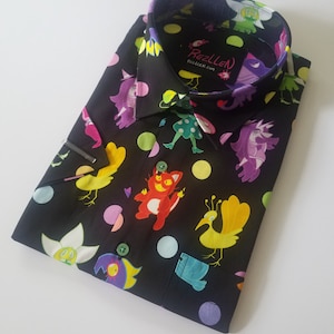Pode incluir: Uma camisa preta de manga curta com botões e uma estampa de desenho animado colorida. A estampa apresenta várias criaturas, incluindo um gato, um pássaro e uma flor. A camisa tem colarinho e um bolso do lado esquerdo. A camisa tem o texto "The Rezzlen" no interior do colarinho.