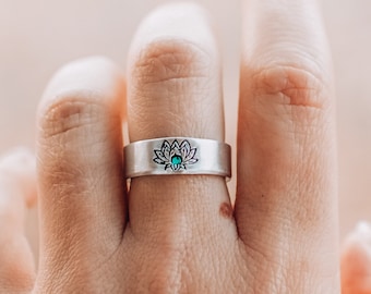May include: A silver band ring featuring a stamped lotus flower design on the front surface. The center of the lotus flower is accented with a small, round, vibrant turquoise-colored stone inlay, creating a distinct focal point.