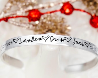 May include: A silver-toned metal cuff bracelet featuring the engraved names Landen, Drew, and Jack, each separated by a small heart symbol. The bracelet has a smooth, polished finish and an open-ended design for easy wear on the wrist.