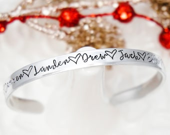 May include: A silver-toned metal cuff bracelet featuring engraved black script text that reads Lauren, Landen, Drew, and Jack, with small heart symbols interspersed between each of the names along the outer surface of the band.