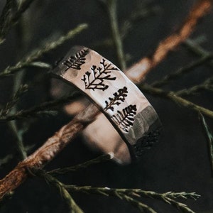 May include: A silver-toned metal ring featuring a band engraved with a repeating pattern of dark, detailed pine tree silhouettes. The ring is positioned on a thin, textured brown branch amidst dark green evergreen needles.