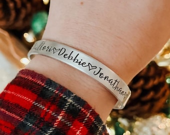 May include: A silver-toned metal cuff bracelet featuring the engraved names Mallory, Debbie, and Jonathan, each separated by a small heart symbol. The bracelet has a smooth, polished finish and is worn on a wrist.