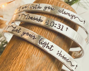 May include: Three silver-toned metal cuff bracelets stacked on a wooden stand. Each bracelet features engraved black text: I am with you always, Isaiah 40:31, and Let your light shine, all accented with small cross symbols at the ends.
