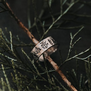 May include: A silver ring with a delicate design of leaves around the band. The ring is resting on a branch of a green evergreen tree.