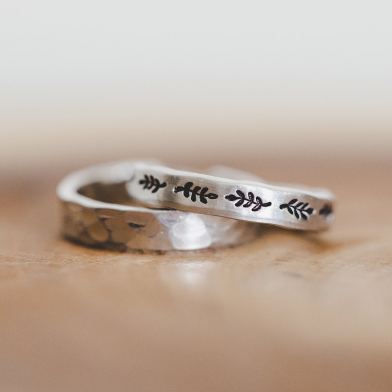 May include: Close-up of two silver rings. One ring has a hammered texture, and the other has a repeating leaf pattern. Both rings appear to be made of metal and have a simple, minimalist design. The rings are stacked on top of each other, showcasing both textures and styles.