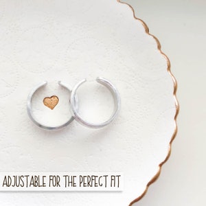Puede incluir: Dos anillos ajustables de plata con un peque&ntilde;o coraz&oacute;n de oro en uno de los anillos. Los anillos est&aacute;n en un plato blanco con un borde dorado. "ADJUSTABLE FOR THE PERFECT FIT" est&aacute; escrito debajo de los anillos.