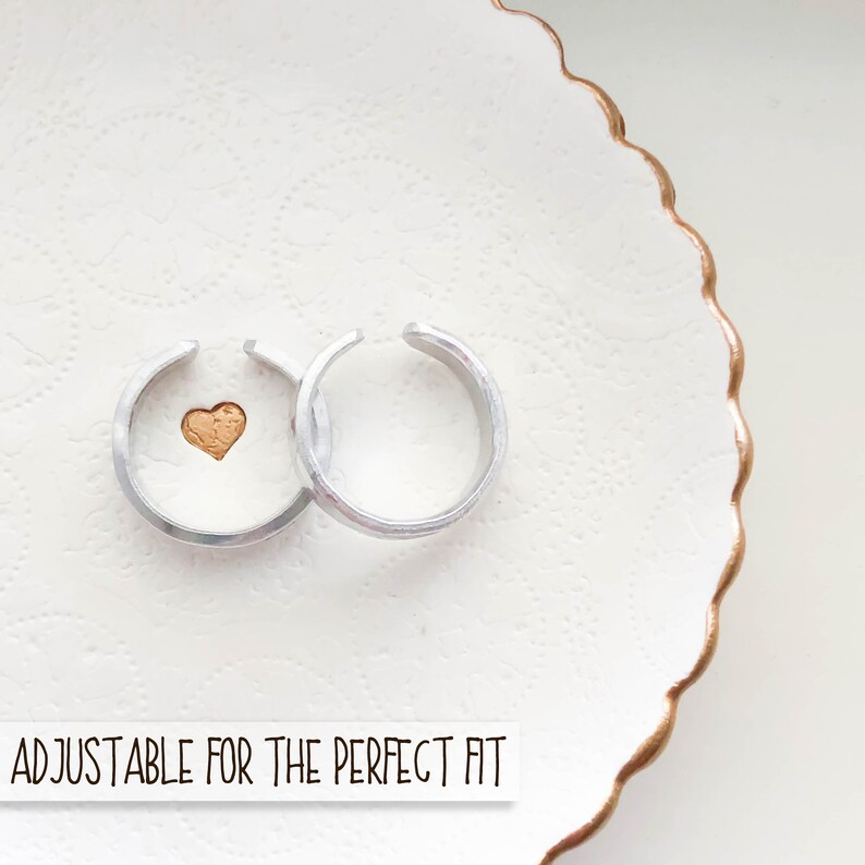Puede incluir: Dos anillos ajustables de plata con un peque&ntilde;o coraz&oacute;n de oro en uno de los anillos. Los anillos est&aacute;n en un plato blanco con un borde dorado. "ADJUSTABLE FOR THE PERFECT FIT" est&aacute; escrito debajo de los anillos.