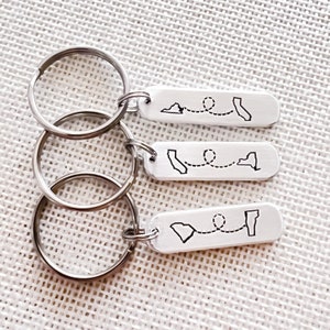 May include: Three silver keychains with engraved outlines of states and a dotted line connecting them. The states are California, New York, and South Carolina.