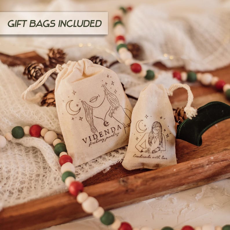 Puede incluir: Dos peque&ntilde;as bolsas de regalo de tela blanca con cierre de cord&oacute;n. Las bolsas tienen un dise&ntilde;o impreso de una mujer con una luna creciente y estrellas. Una bolsa dice "Videnda Jewelry" y la otra dice "Hecho a mano con amor".