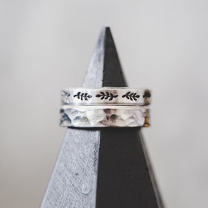 Puede incluir: Dos anillos de plata apilados en un soporte triangular negro y gris. El anillo superior tiene un delicado dise&ntilde;o de hojas. El anillo inferior tiene una textura martillada.