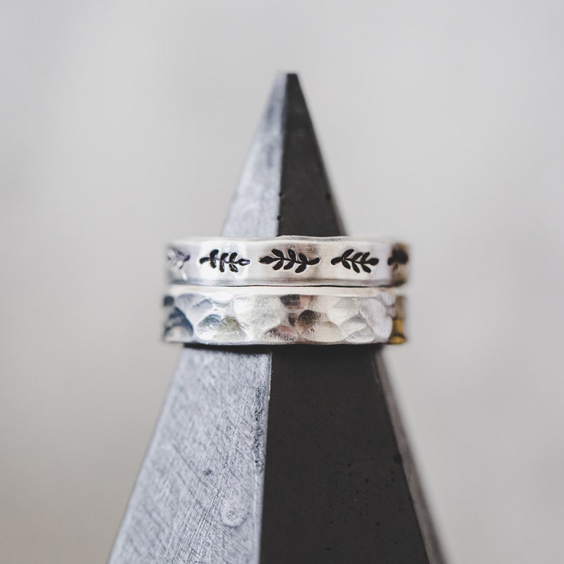 Puede incluir: Dos anillos de plata apilados en un soporte triangular negro y gris. El anillo superior tiene un delicado dise&ntilde;o de hojas. El anillo inferior tiene una textura martillada.