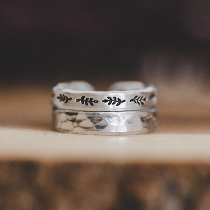 Puede incluir: Dos anillos de plata apilados uno encima del otro. El anillo superior tiene un acabado martillado y el anillo inferior tiene una fila de peque&ntilde;os dise&ntilde;os de hojas.