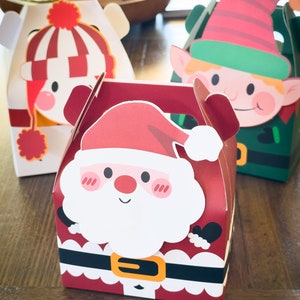 May include: Three festive Christmas gift boxes with different character designs. The boxes are red, white, and green with cartoon images of Santa Claus, an elf, and a snowman. The boxes have a handle on top and are perfect for holding small gifts.