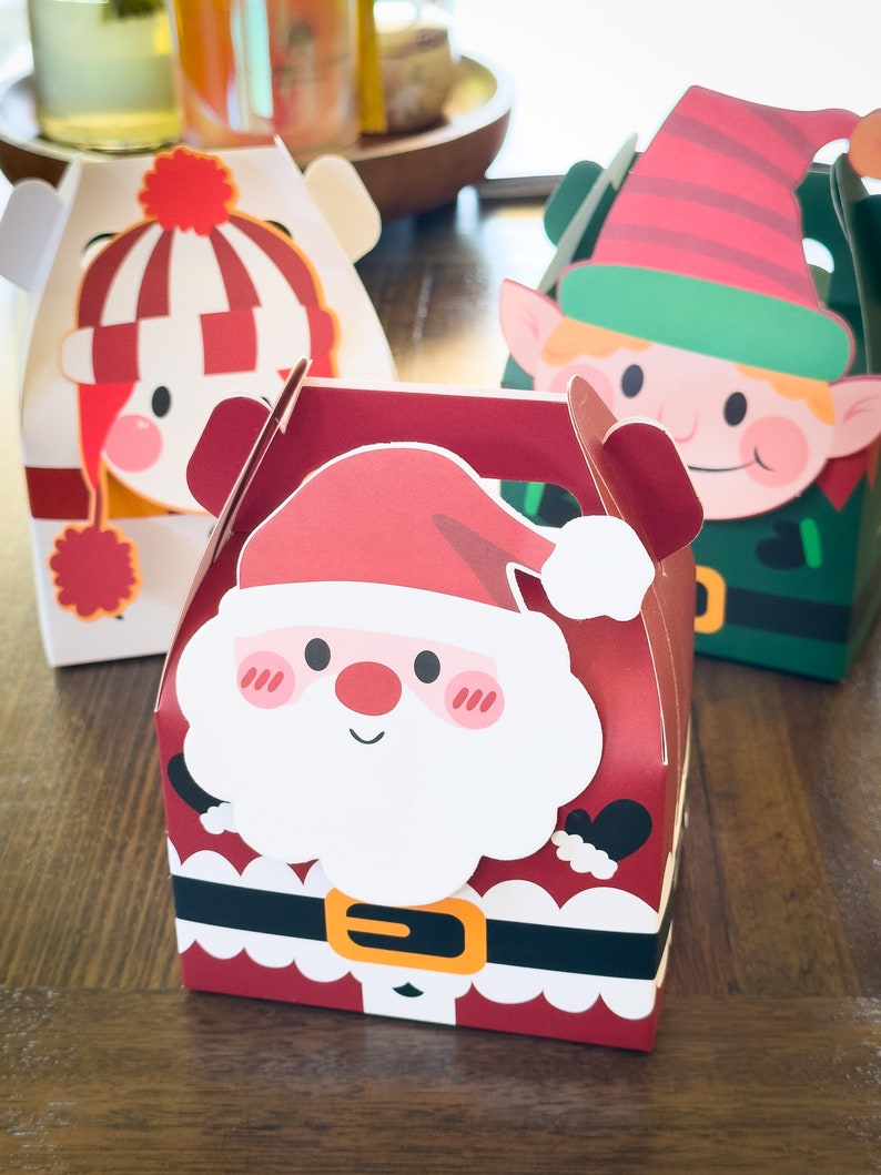 May include: Three festive Christmas gift boxes with different character designs. The boxes are red, white, and green with cartoon images of Santa Claus, an elf, and a snowman. The boxes have a handle on top and are perfect for holding small gifts.