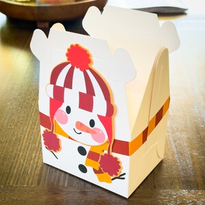 May include: A white cardboard box with a snowman design. The snowman is wearing a red and white striped hat with a red pom-pom, a red scarf, and a yellow scarf. The box has a red and orange striped ribbon.