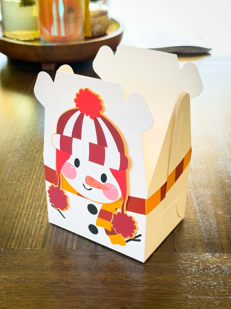 May include: A white cardboard box with a snowman design. The snowman is wearing a red and white striped hat with a red pom-pom, a red scarf, and a yellow scarf. The box has a red and orange striped ribbon.