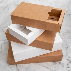 May include: Four cardboard boxes stacked on top of each other. The top box is brown with a cut-out window. The second box is white with a cut-out window. The third box is brown and the bottom box is brown.