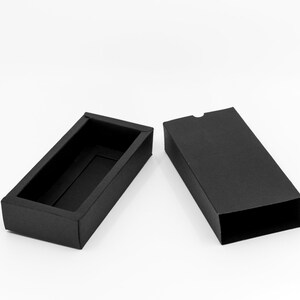 May include: A black rectangular box with a lid, open to reveal an empty interior. The box is made of cardboard and has a smooth, matte finish.