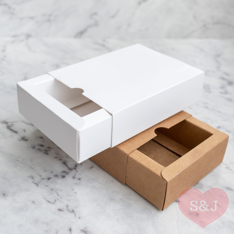 May include: Two boxes, one white and one brown, are stacked on top of each other. The white box has a sliding lid and the brown box has a cutout in the top. Both boxes have a rectangular shape.