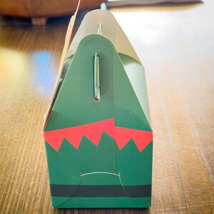 May include: A green and red gift box with a black belt and a white handle. The box is decorated with a festive elf design.