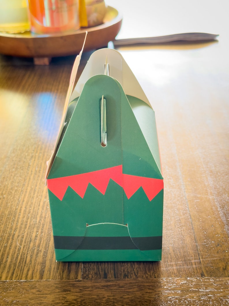 May include: A green and red gift box with a black belt and a white handle. The box is decorated with a festive elf design.