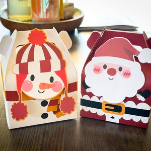 May include: Two festive gift boxes with a snowman and Santa Claus design. The snowman box is white with red and orange accents, while the Santa box is red with white and black accents.