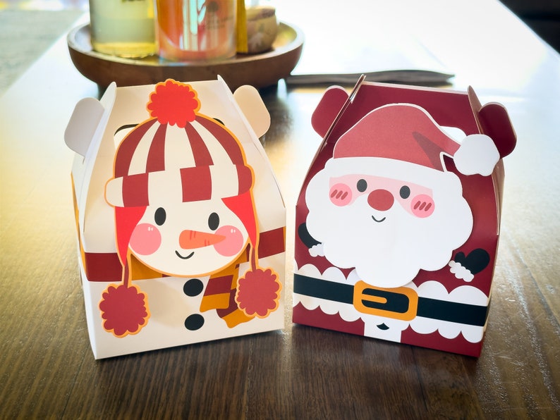 May include: Two festive gift boxes with a snowman and Santa Claus design. The snowman box is white with red and orange accents, while the Santa box is red with white and black accents.