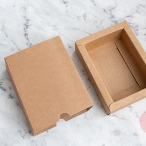 May include: Two brown cardboard boxes, one with a lid and one without, on a white marble surface. The box with the lid is open, revealing the empty interior.