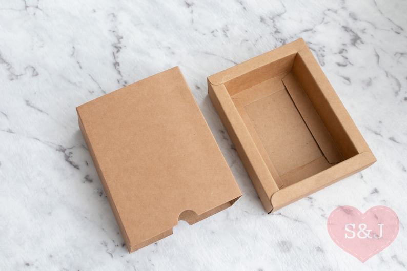 May include: Two brown cardboard boxes, one with a lid and one without, on a white marble surface. The box with the lid is open, revealing the empty interior.