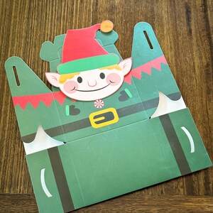 May include: A green and red paper box shaped like an elf with a white candy cane and a yellow belt. The elf has a red hat, green collar, and a smiling face.