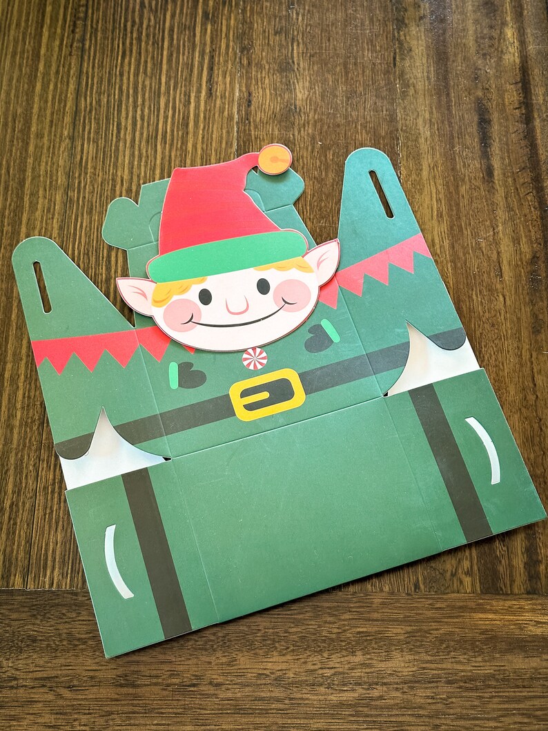 May include: A green and red paper box shaped like an elf with a white candy cane and a yellow belt. The elf has a red hat, green collar, and a smiling face.