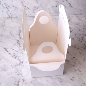 K&ouml;nnte beinhalten: Wei&szlig;e Pappbox mit zwei ausgeschnittenen Griffen. Die Box ist ge&ouml;ffnet und das Innere ist sichtbar. Die Box ist zum Verpacken und Tragen kleiner Gegenst&auml;nde gedacht.